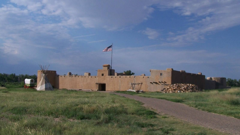 Bent's Old Fort beneath a blue sky, with an American flag flying above the adobe walls.