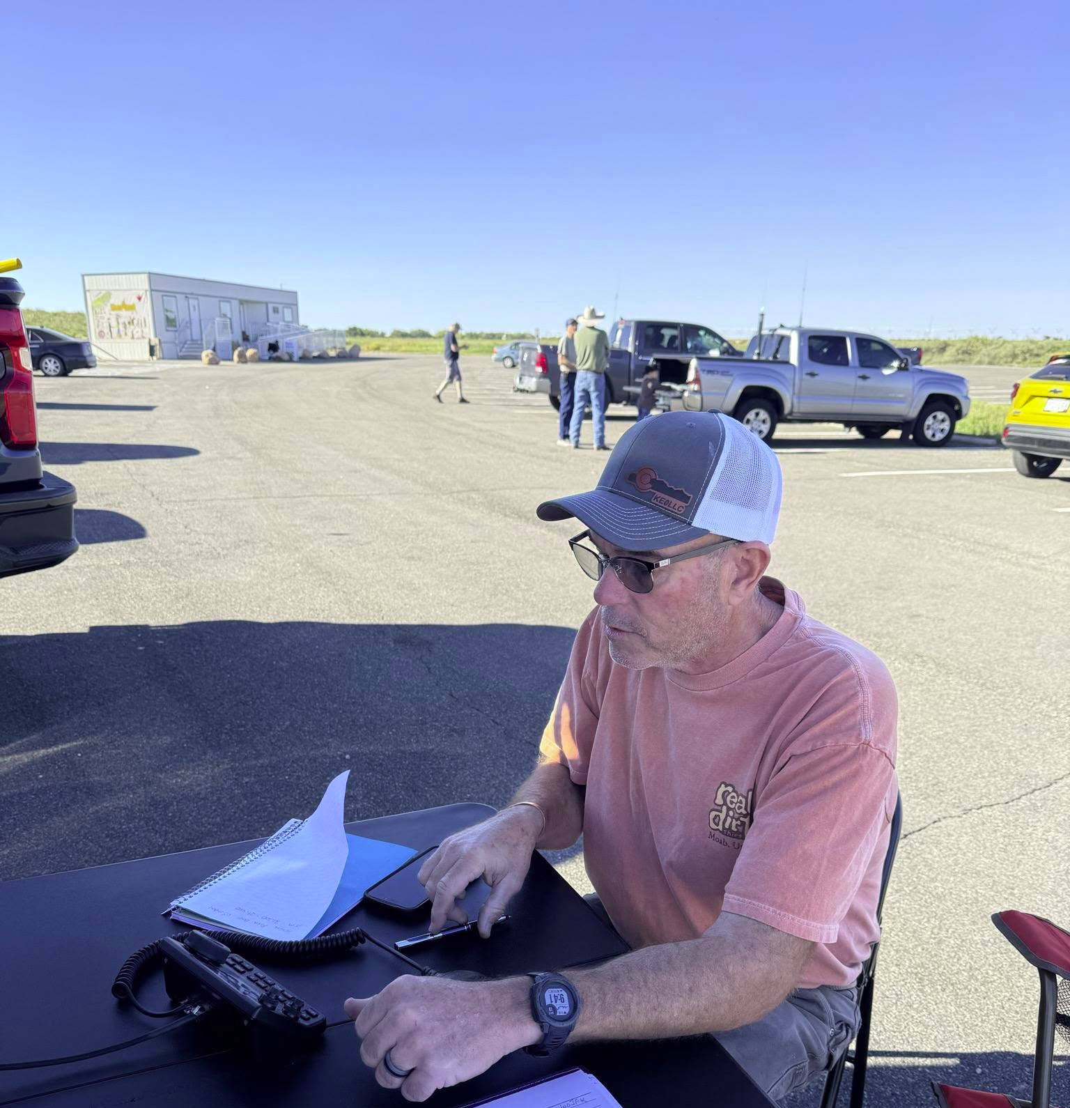 An amateur radio operator sits at an outdoor table with a transceiver and microphone.