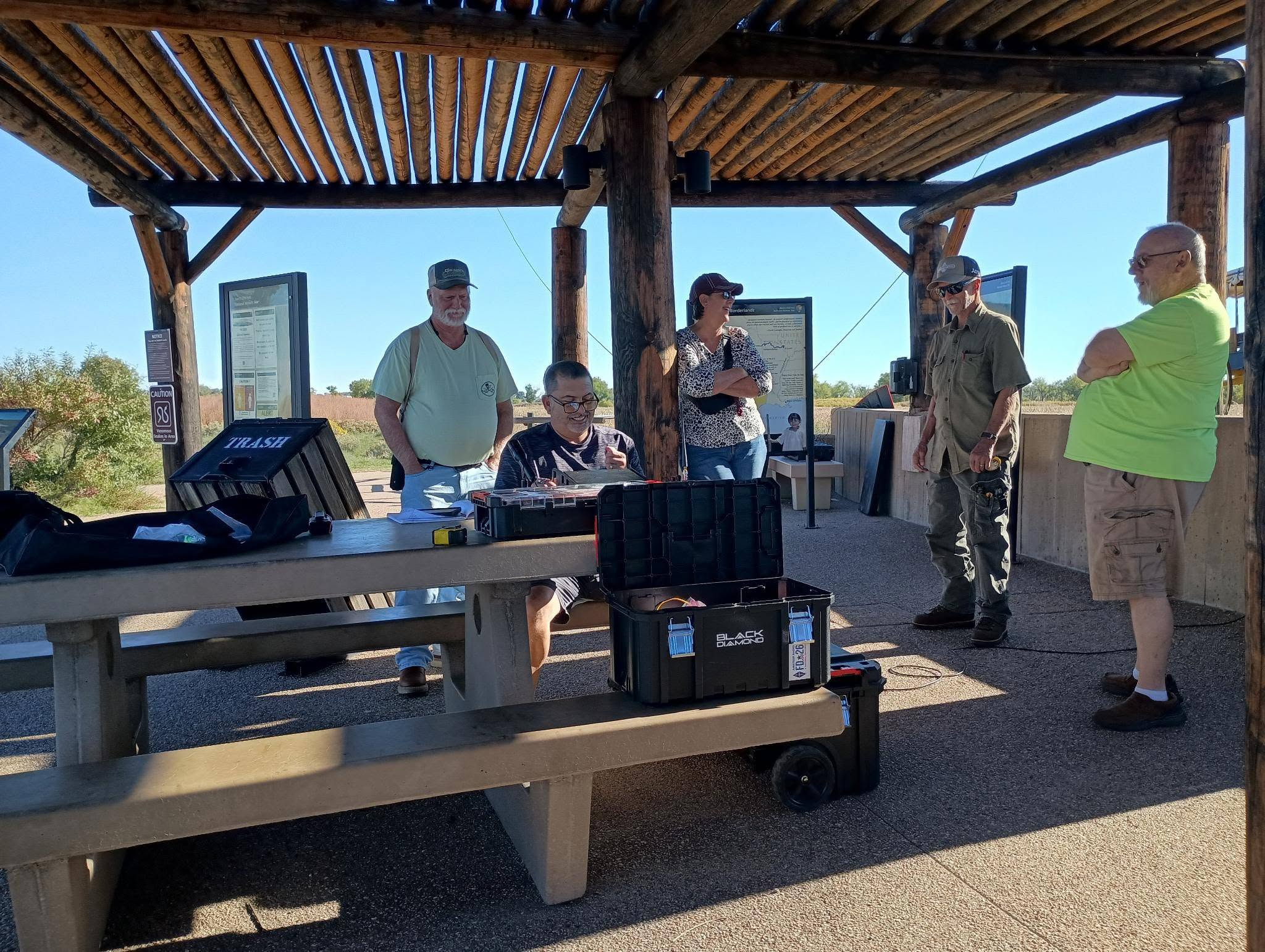 Participants gather around a portable amateur radio station and equipment cases beneath a wooden shelter.